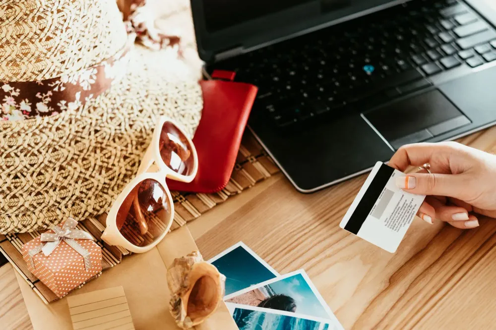 Table filled with travel gear, sunglasses, a laptop, and a woman's hand holding a credit card